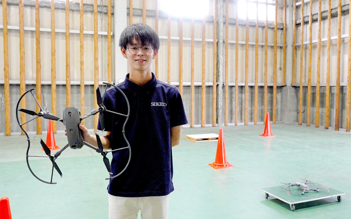 屋内飛行場で、プロペラガードを付けたドローンを手にする講習スタッフ。三角コーンと離着陸パッドが並ぶ