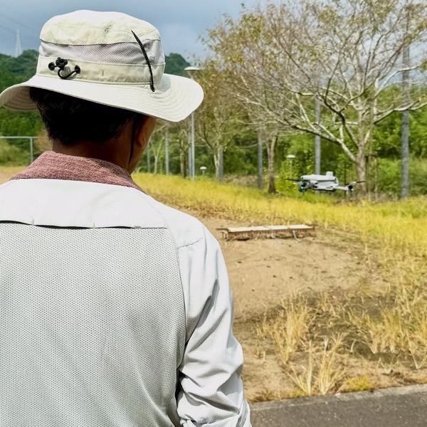 帽子をかぶったお客様が、草地の広がる現場で「DJI Matrice 4E」の操縦講習を受けている後ろ姿