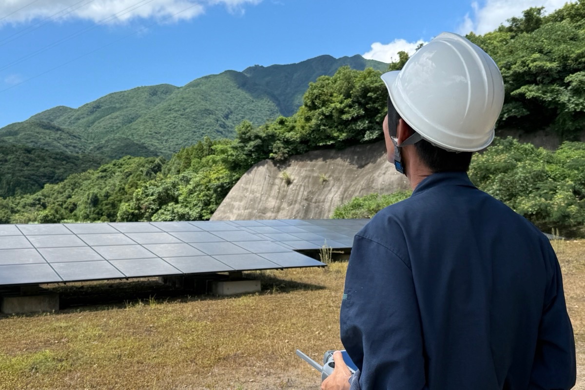 山あいの太陽光発電所で、ヘルメットを着けた作業員が送信機を手にドローンを操縦している